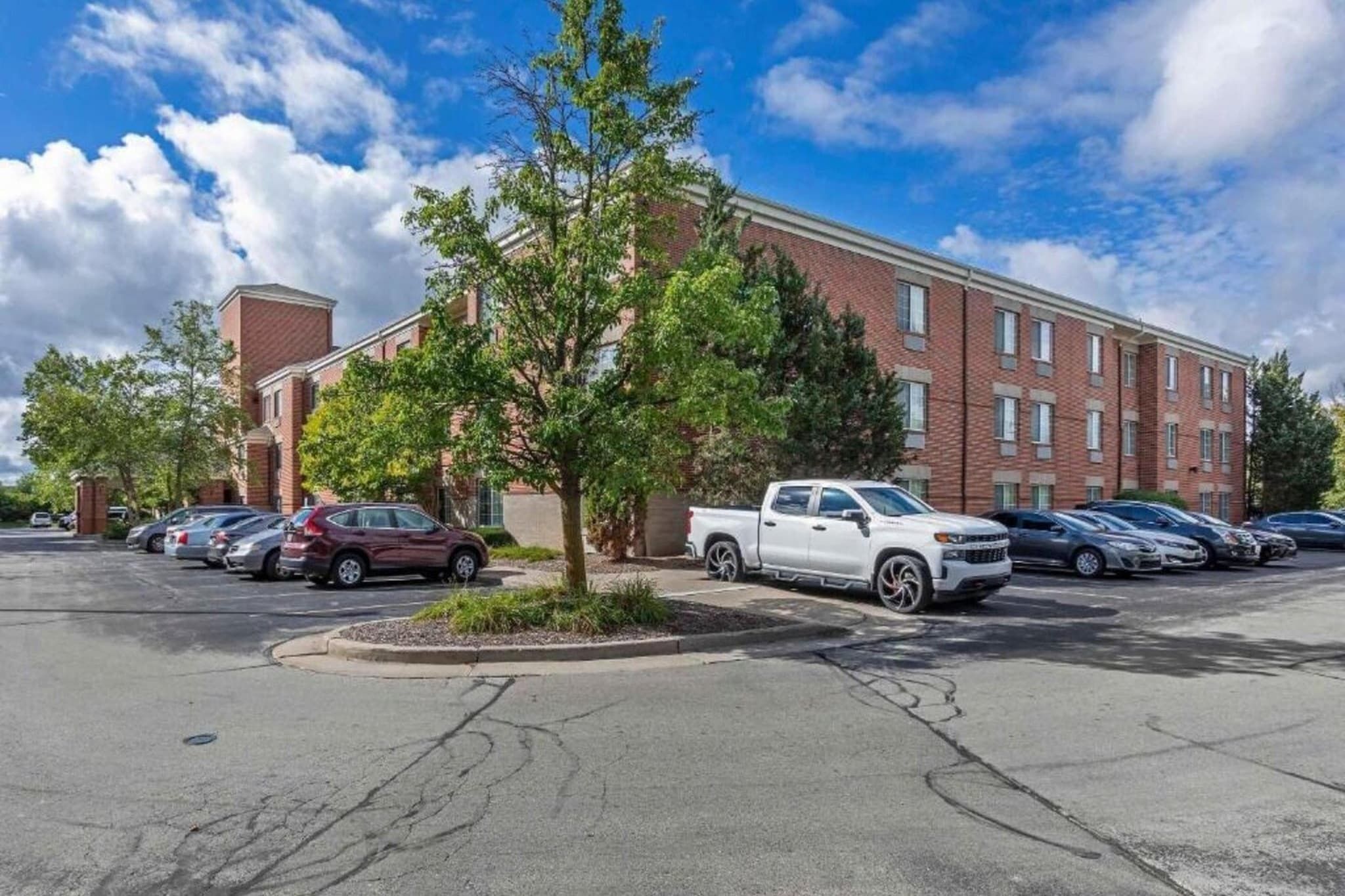 A parking lot with several cars, a white pickup truck, and a brick building with multiple windows.
