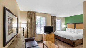Habitación de hotel con una cama, un televisor sobre una cómoda de madera, un sofá y dos ventanas con cortinas.