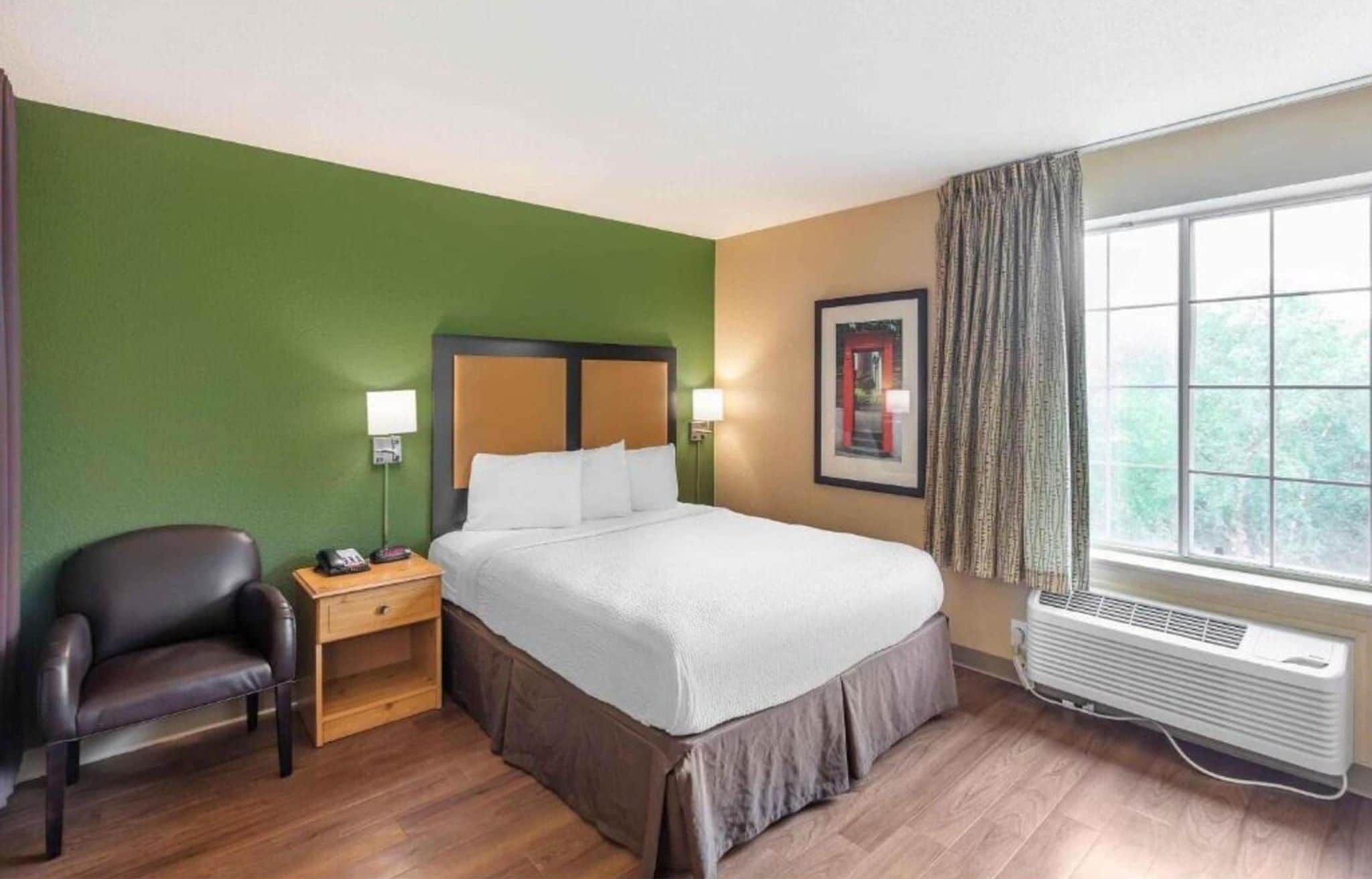 A hotel room with a bed, a chair, a nightstand, a wall-mounted lamp, a framed picture, and a window with curtains.