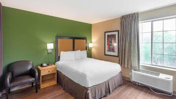 A hotel room with a bed, a chair, a nightstand, a wall-mounted lamp, a framed picture, and a window with curtains.