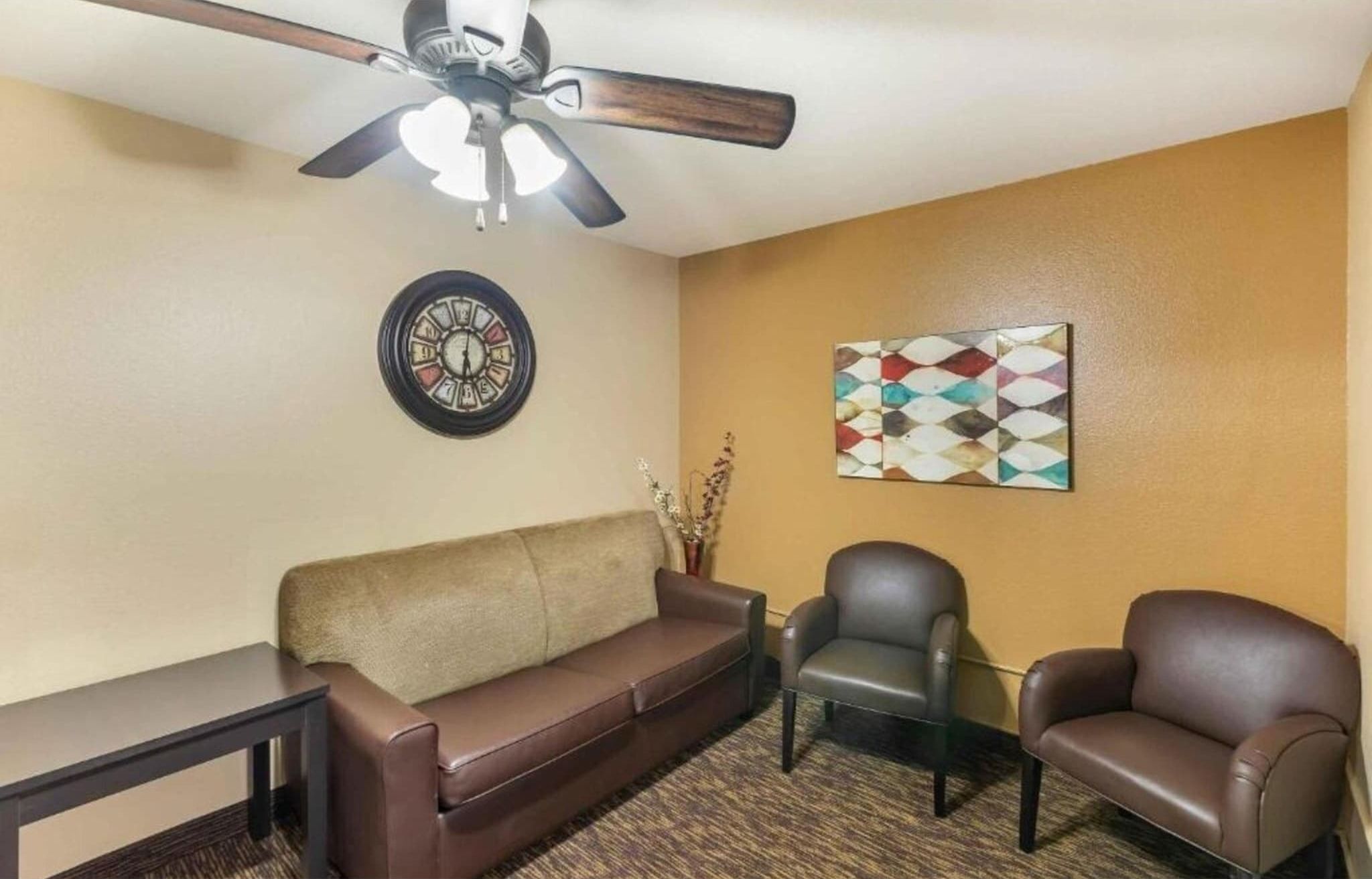 A room with a ceiling fan, a clock, a couch, two chairs, and a decorative wall piece.