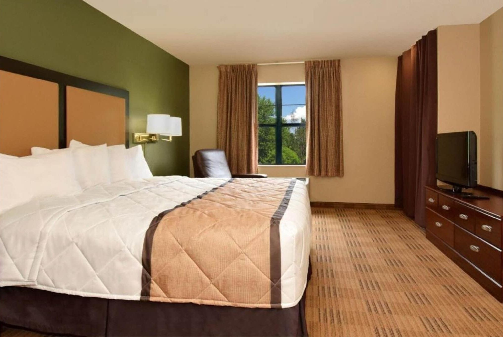 A hotel room with a large bed, a television, a desk with drawers, a chair, and a window with curtains.