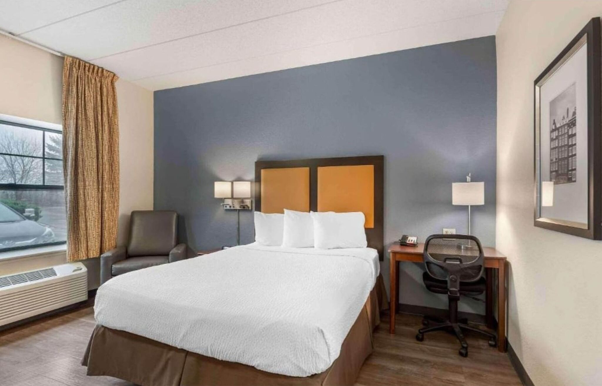 A hotel room with a bed, a desk, a chair, a lamp, and a window with curtains.