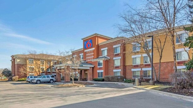 A multi-story hotel with a prominent logo on the facade, a parking lot with several cars, and a clear sky. - 1 Queen Bed|ADA/RI Shower/Smk/Kitchen (Vernon Hills)