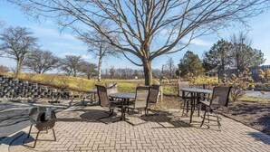An outdoor patio with brick flooring, metal tables, and chairs, surrounded by trees and a grassy area. - 1 Queen Bed|ADA/RI Shower/Smk/Kitchen (Vernon Hills)