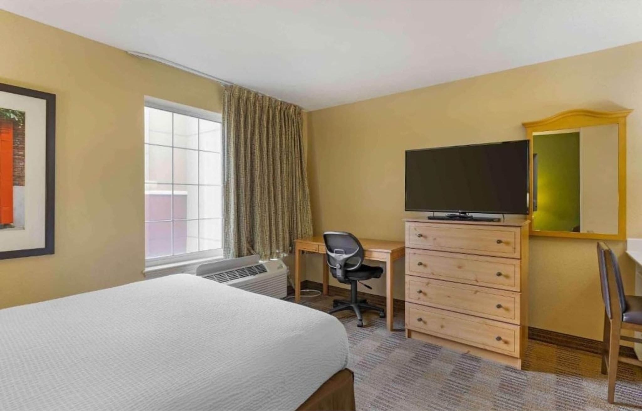 A hotel room with a bed, a desk, a chair, a dresser, a television, and a window with curtains.