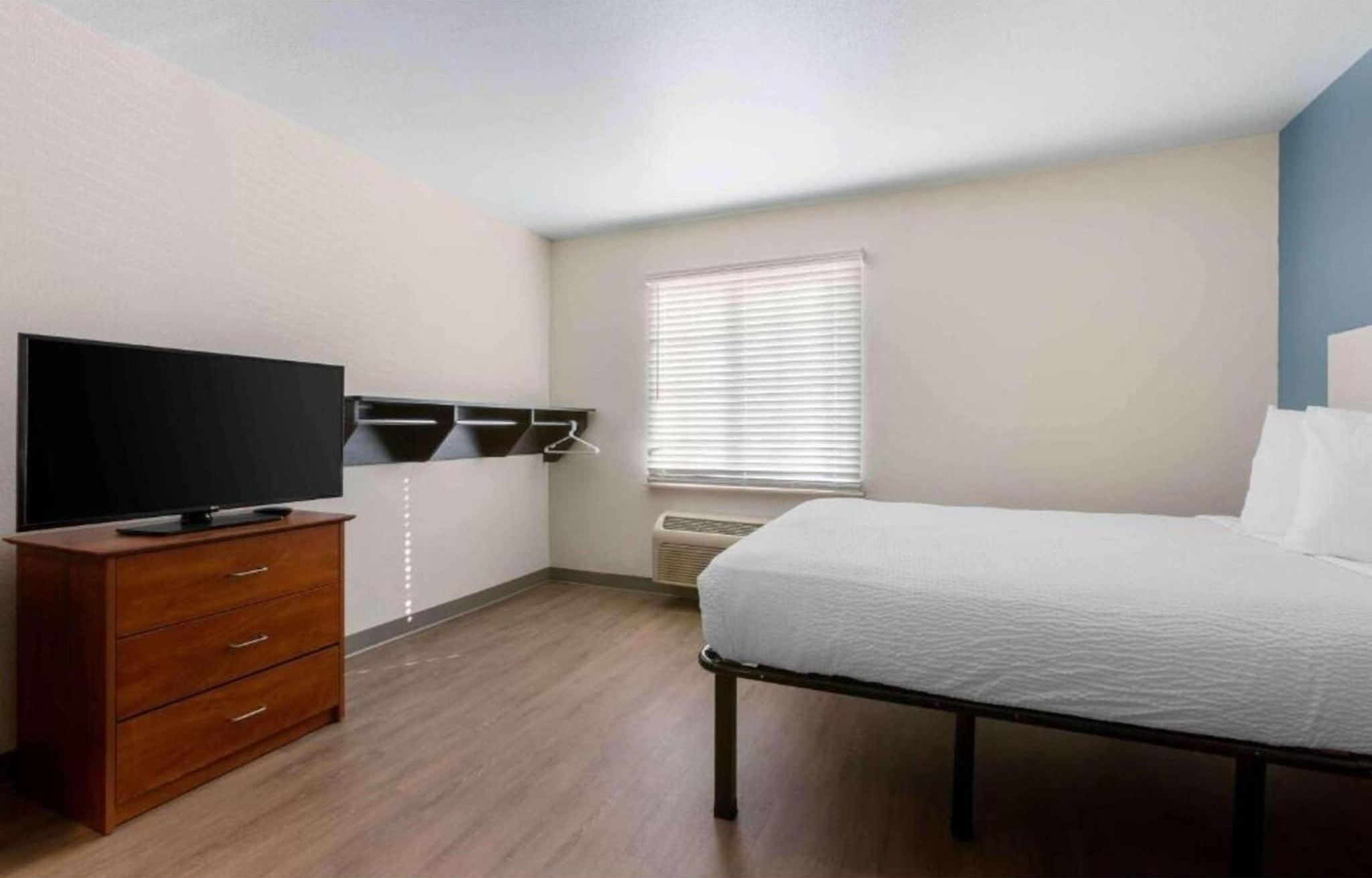 A bedroom with a bed, a TV on a wooden dresser, a window with blinds, and an air conditioning unit.