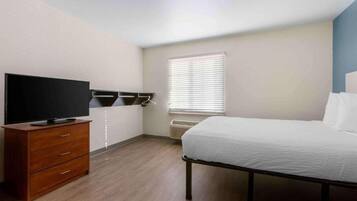 A bedroom with a bed, a TV on a wooden dresser, a window with blinds, and an air conditioning unit.