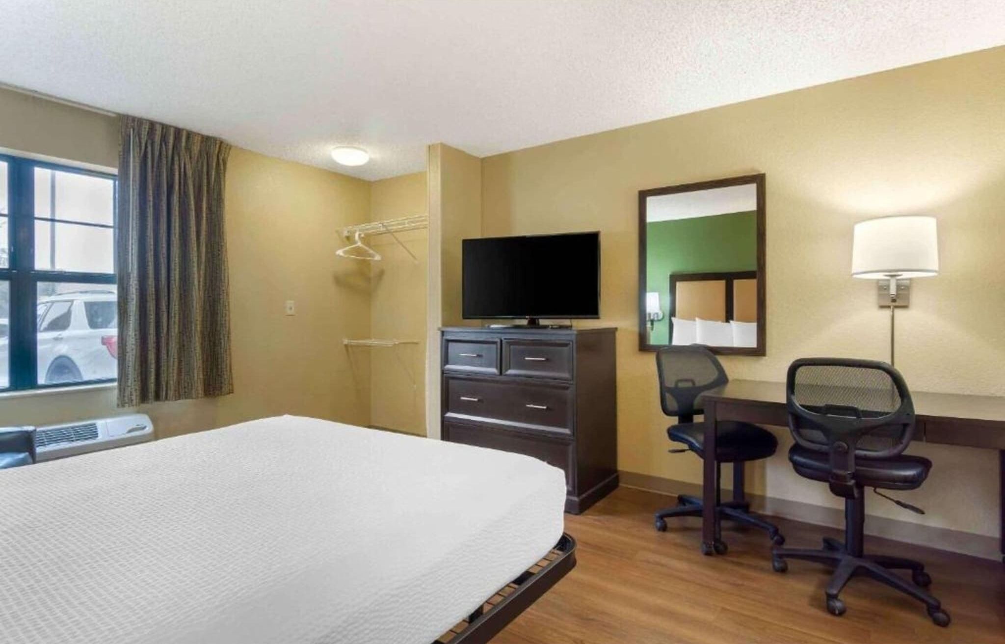 A hotel room with a bed, a desk with a chair, a television, a mirror, and a window with curtains.