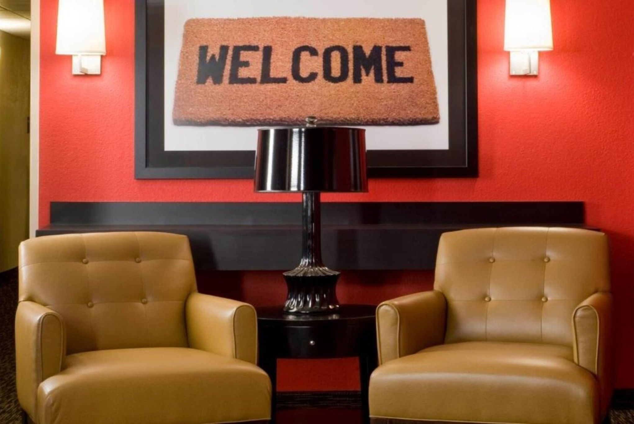 Een hotel lobby met twee leren fauteuils, een vloermat, een bord met welkomstboodschap en een lamp op een bijzettafeltje.