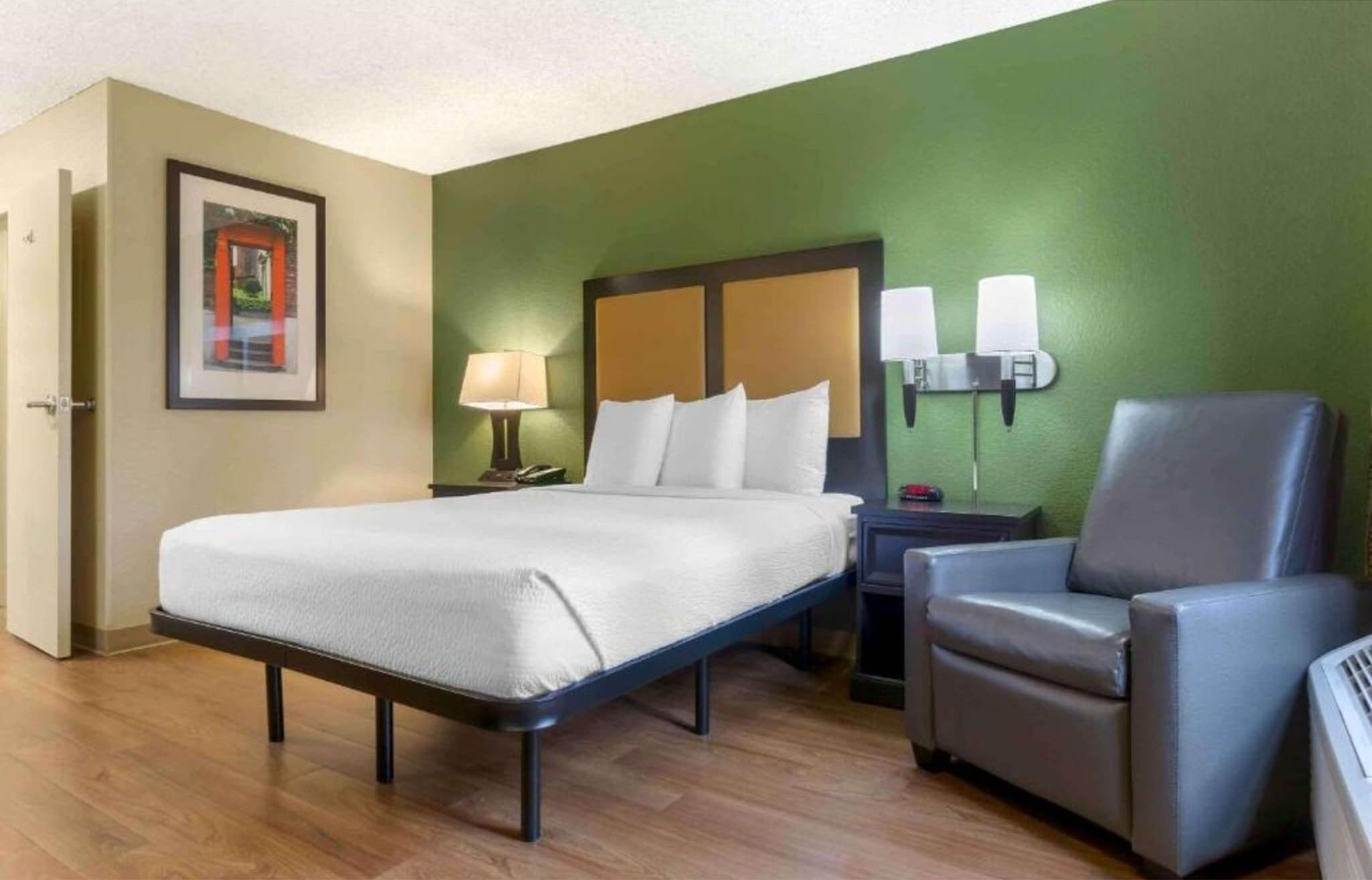 A hotel room with a bed, a chair, a nightstand, a lamp, and a wall-mounted light fixture.