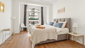 Ein modernes Schlafzimmer mit einem großen Bett, einem Nachttisch und Blick auf einen Balkon mit Geländer.