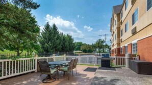 Eine Terrasse mit Gartenmöbeln, ein Gebäude mit vielen Fenstern und ein von Bäumen gesäumter, umzäunter Bereich.