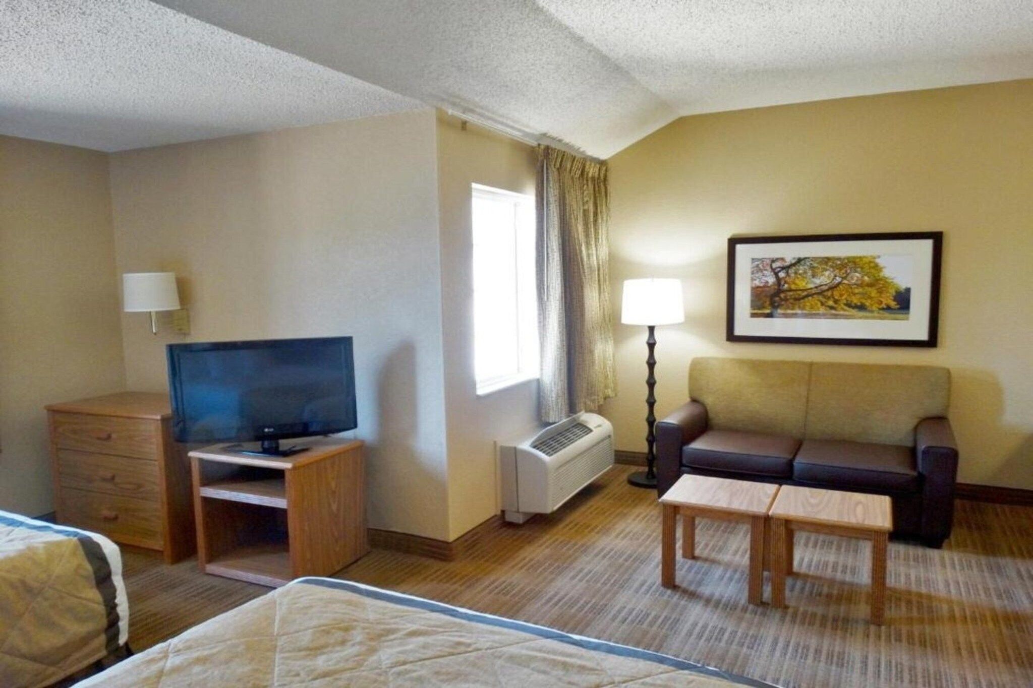 A hotel room with a bed, a TV on a stand, a sofa, a table, a lamp, and a framed picture on the wall.
