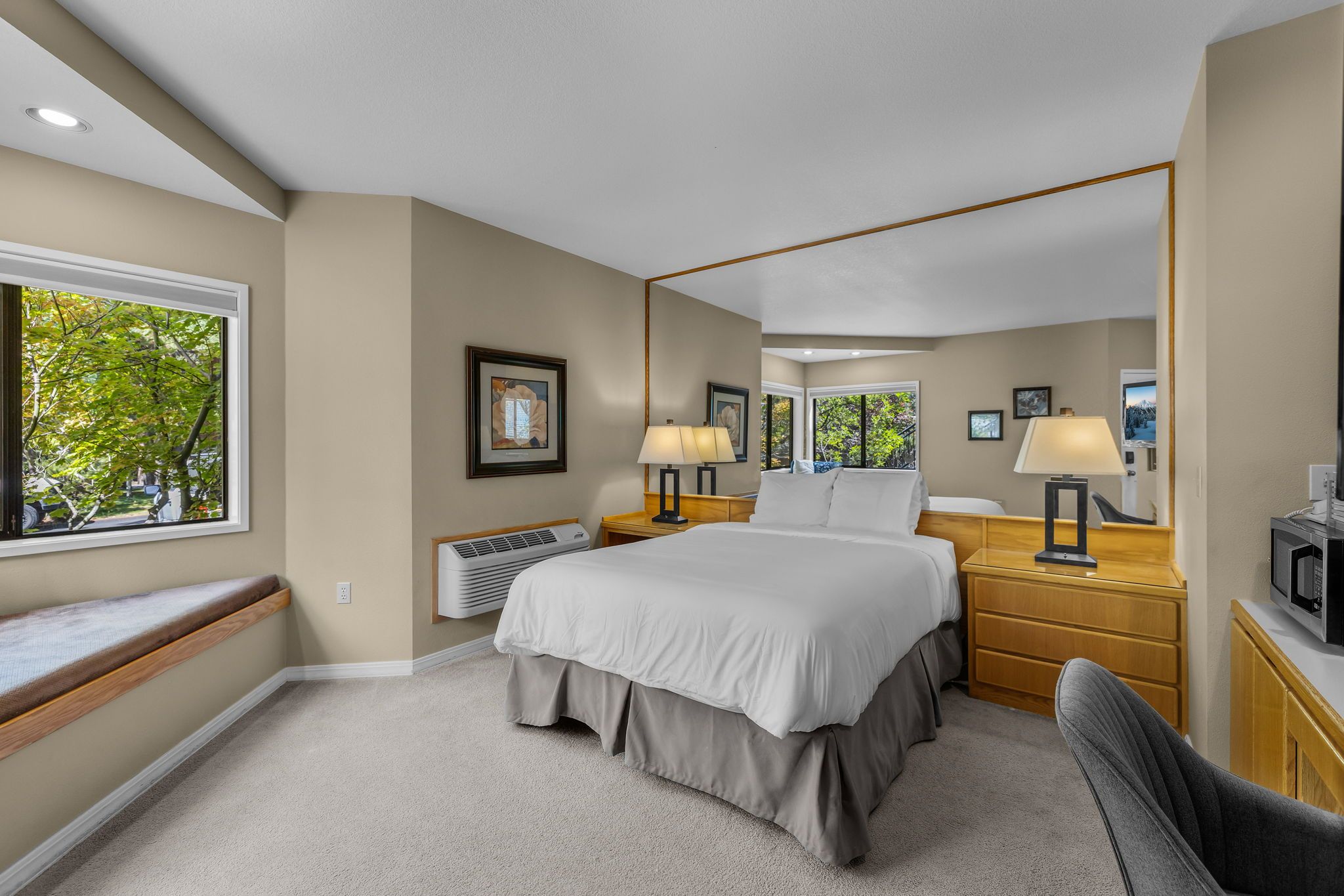 A hotel room with a large bed, two bedside tables with lamps, a built-in air conditioner, a window with a view of trees, and a small desk with a microwave.