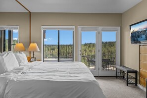 Ein Schlafzimmer mit einem großen Bett, zwei Nachttischlampen, einem Fernseher und einem Balkon mit Blick auf Bäume und einen Berg.