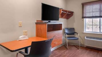 A room with a wooden desk, a chair, a television mounted on a cabinet, a window with blinds, and a wall-mounted phone.