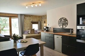 A compact kitchen with a stone wall, a dining table with chairs, a coffee machine, and a modern oven. - Holiday home with 1 bedroom (SAINT-LATTIER)