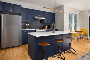 A modern kitchen with dark blue cabinets, a stainless steel refrigerator, a coffee maker, and a gas stove. There are three wooden stools at the island. - Accessible 1BR Apartment Fishtown - StayCozy (Philadelphia)