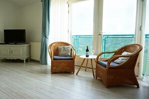 Ein Zimmer mit zwei Korbstühlen, einem kleinen runden Tisch mit Flasche und Glas, einem Fernseher auf einer weißen Kommode und Blick auf einen Balkon mit blauer Brüstung.