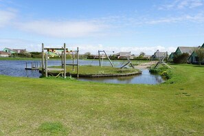 Ein Spielplatz mit Schaukeln und einem kleinen Teich, umgeben von Gras und Häusern.