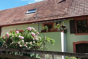 Ein Gebäude mit einem Ziegeldach, einem Balkon mit hängenden Blumenkörben und einem großen, blühenden Strauch im Vordergrund.