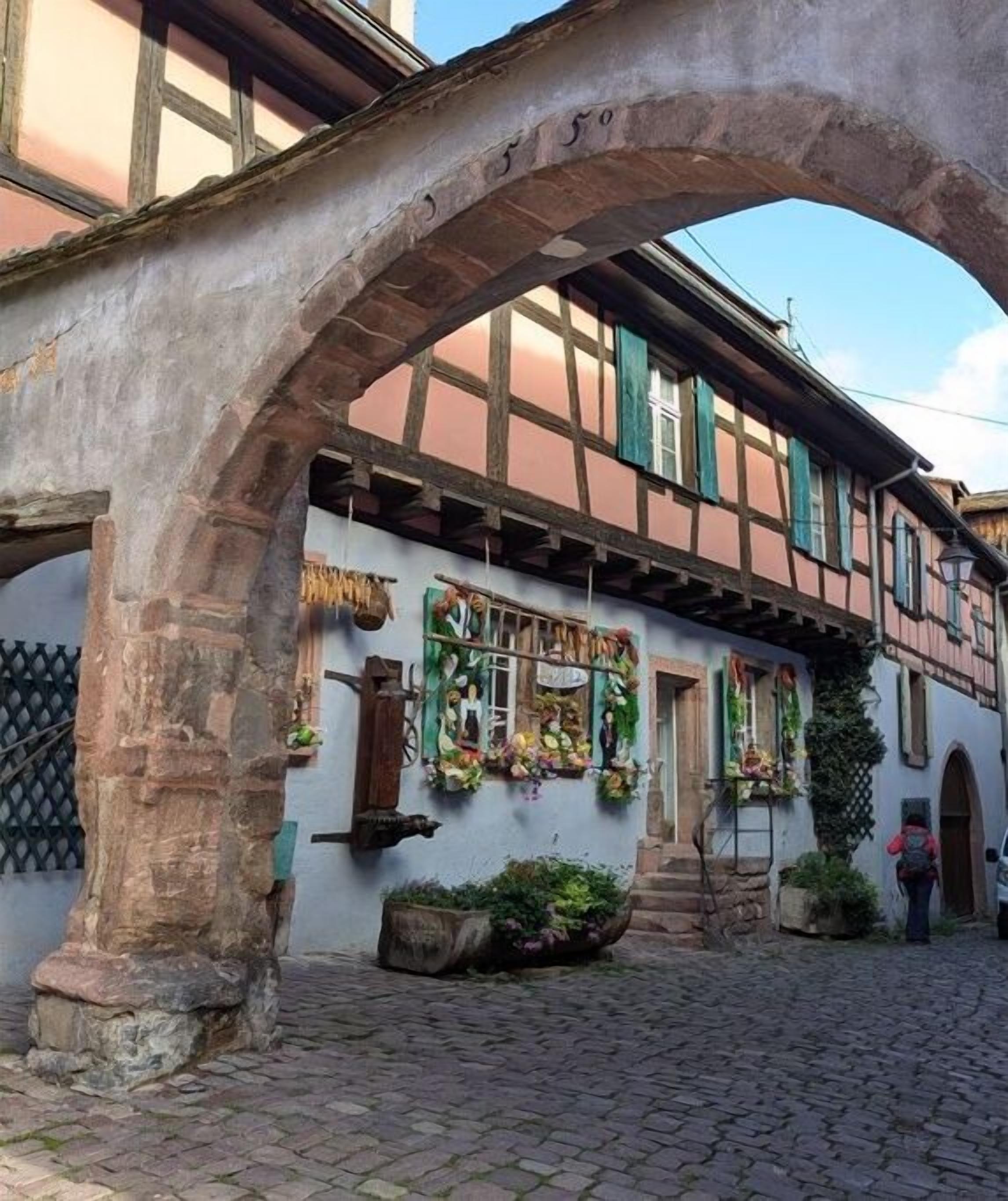 Un bâtiment traditionnel à colombages, doté d’une arche en pierre, d’une rue pavée et de jardinières suspendues.