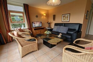 A living room with a black sofa, wicker chairs, a coffee table, a TV, and a window with curtains. - Holiday home with 1 bedroom (Medemblik)