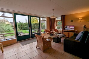 Un salon comprenant un canapé, des fauteuils, une table basse, un téléviseur et donnant sur un jardin grâce à de grandes fenêtres.