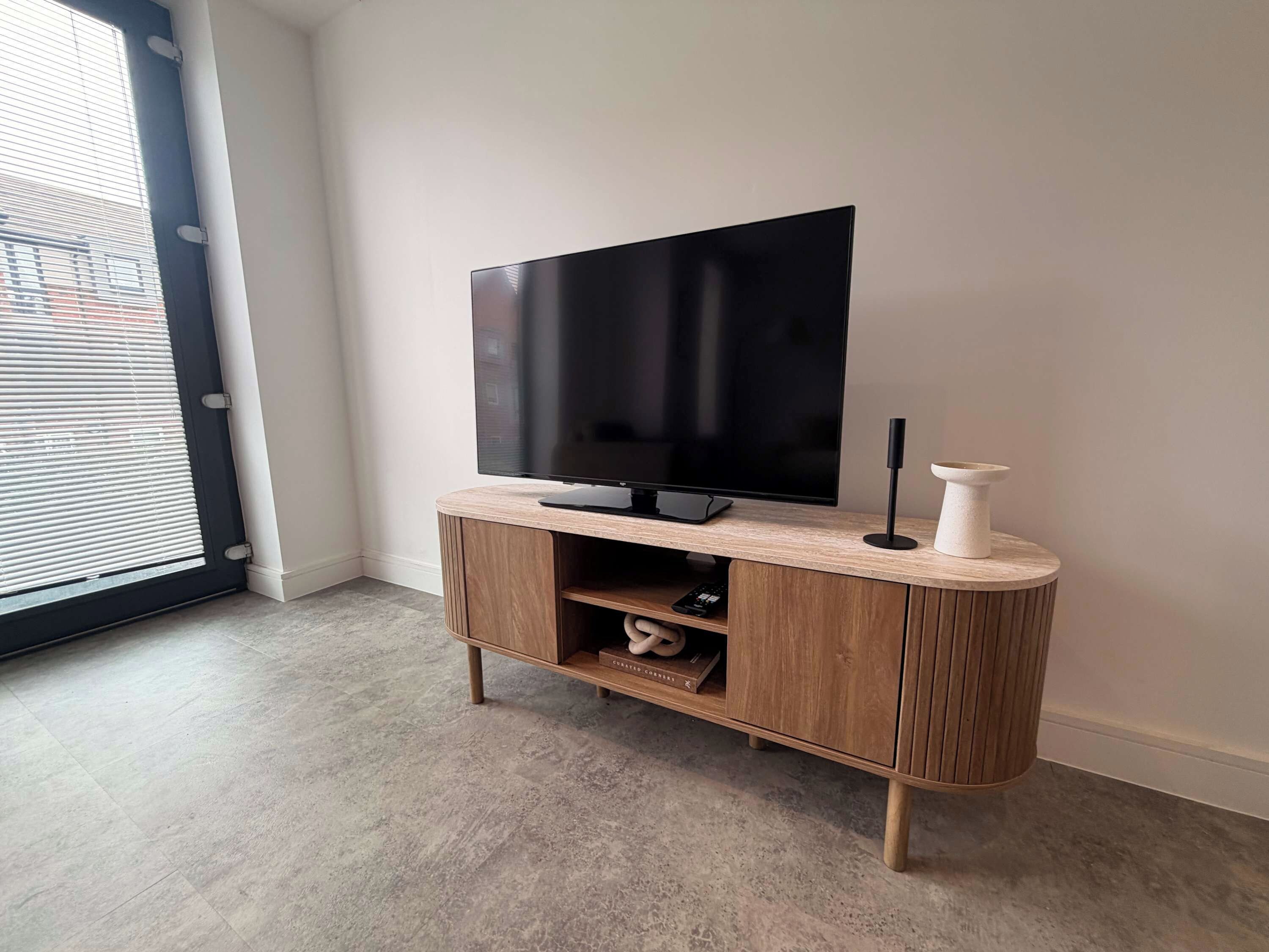 A modern living room with a flat-screen TV on a wooden stand, a remote control, and a white vase.