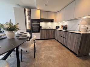 A modern kitchen with wooden cabinets, a built-in oven, and a dining area with a table and chairs. - Stylish 2-Bed Poulton Stay | Parking & Self Check-In (Lancashire)