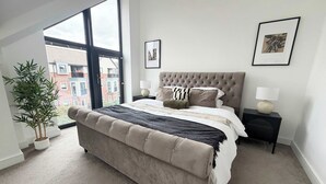 Een slaapkamer met een groot bed, een kamerplant, een raam met uitzicht op gebouwen en een foto in een lijst aan de muur.