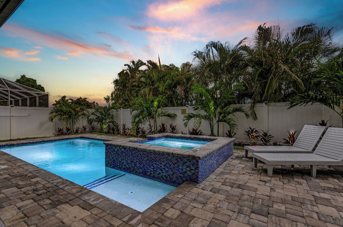 A swimming pool with a tiled edge, surrounded by a brick patio. There are lounge chairs and a small hot tub.