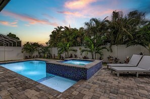 Ein Schwimmbecken mit einer gefliesten Kante, umgeben von einer Ziegelterrasse. Liegestühle und ein kleines Whirlpool sind vorhanden.
