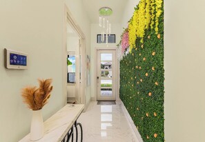 A hallway with a wall-mounted thermostat, a door with a glass panel, and a decorative wall with green foliage and yellow flowers. - New! Wave Chaser • New Luxury Build • Heated Salt Pool/Spa • 1 Mile to Vanderbilt Beach! (Naples)