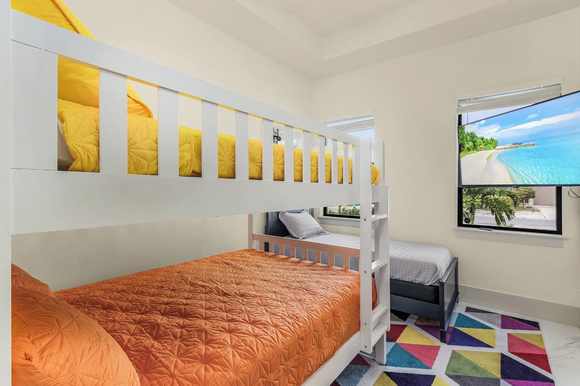 A bunk bed with yellow pillows, a single bed with an orange quilt, a small desk, and a window with a beach view.