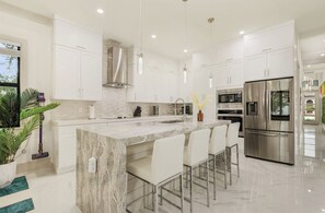 A modern kitchen with white cabinets, stainless steel appliances, and a marble island with bar stools. - New! Wave Chaser • New Luxury Build • Heated Salt Pool/Spa • 1 Mile to Vanderbilt Beach! (Naples)