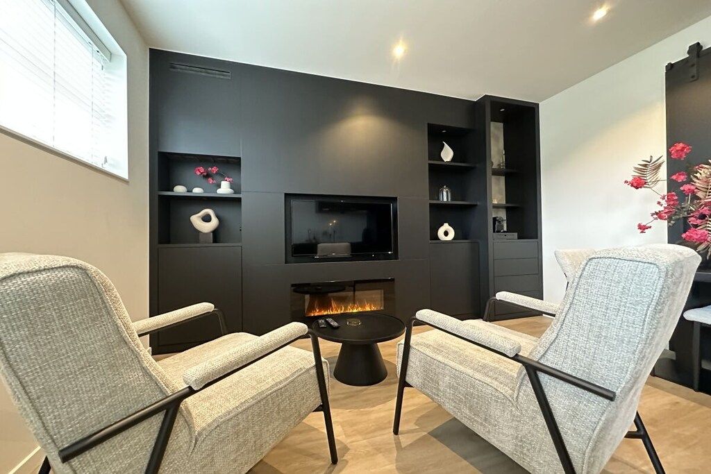 A modern living room with a fireplace, two armchairs, a coffee table, and a flat-screen TV mounted on a dark wall.
