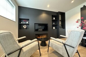 A modern living room with a fireplace, two armchairs, a coffee table, and a flat-screen TV mounted on a dark wall. - Holiday apartment with 1 bedroom (Oudkarspel)