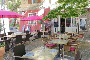 An outdoor café with tables and chairs, some with umbrellas, and a building with a red facade in the background. - with pool in the hinterland of the coast (Flayosc)