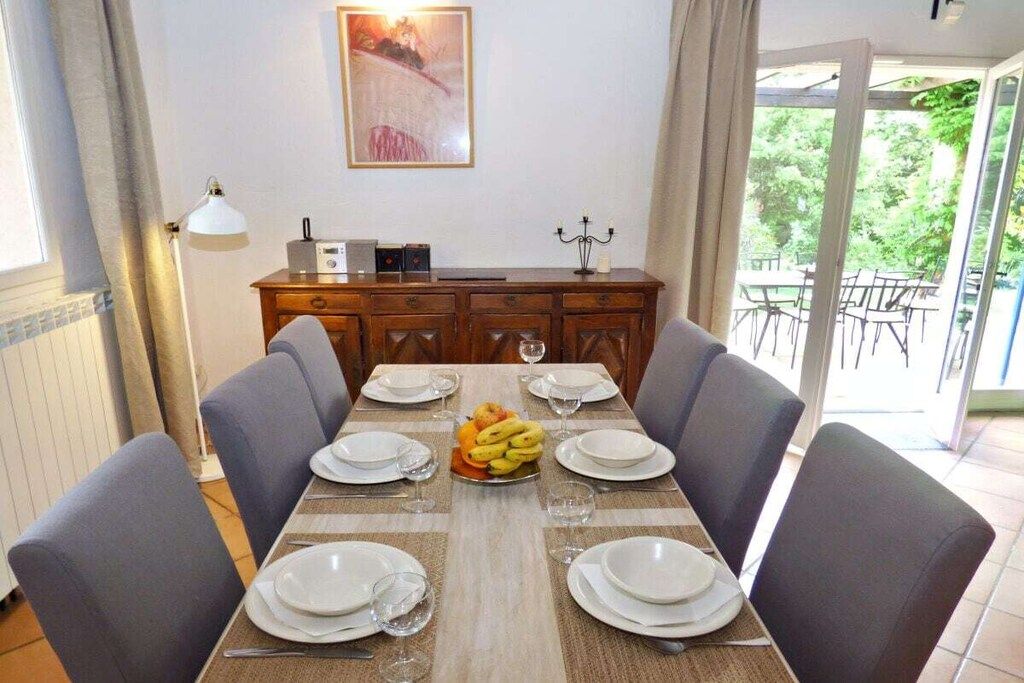 A dining area with a table set for six, a fruit bowl on the table, and a view of the outdoors through a glass door.