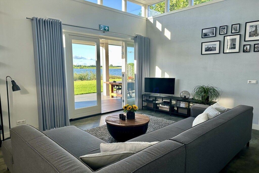 A modern living room with a sofa, a coffee table with flowers, a flat-screen TV, and a view of a lake through a glass door.