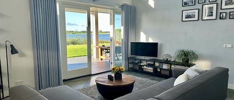 A modern living room with a sofa, a coffee table with flowers, a flat-screen TV, and a view of a lake through a glass door.
