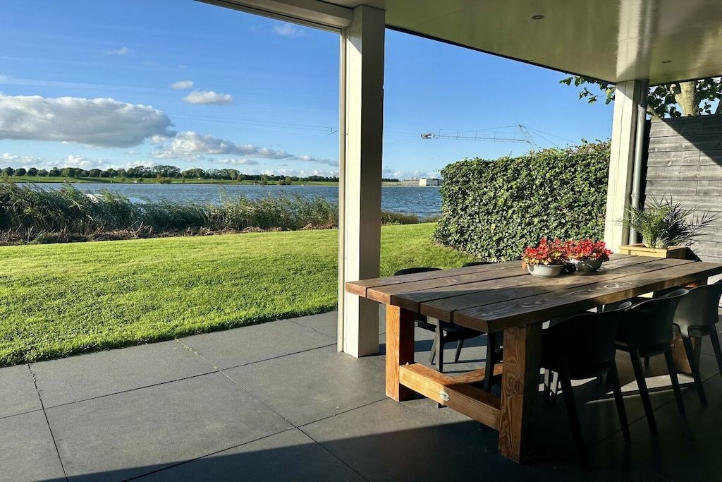 An outdoor patio with a wooden table and chairs, overlooking a body of water and a grassy area.