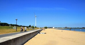 Ein Küstenweg mit einer Betonbarriere, einem Sandstrand und einer Windkraftanlage in der Ferne.