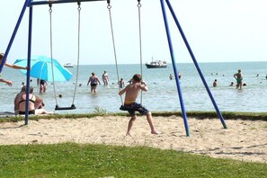 Eine Strandansicht mit einer Schaukel, Personen im Wasser und einem Boot in der Ferne.