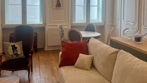 Ein Zimmer mit Holzboden, einem beigen Sofa, einem roten Kissen, einem Holzstuhl, einem Tisch und einer Pendelleuchte.