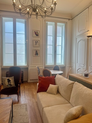 Quarto com piso de madeira, um sofá bege, almofada vermelha, cadeira de madeira, mesa e um candelabro.