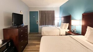 Et hotelværelse med to senge, et fjernsyn på en kommode, et skrivebord med lampe og et vindue med gardiner.