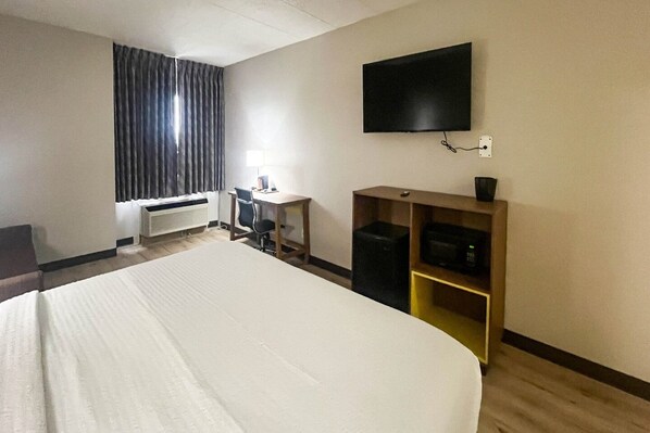 A hotel room with a large bed, a desk, a chair, a flat-screen TV mounted on the wall, and a small shelf with a speaker.